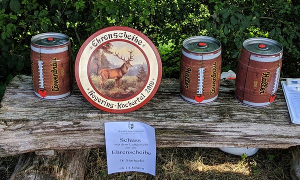 Auf einem Holztisch stehen eine Ehrenscheibe mit einem Rehbock-Motiv und drei Dosen in Fußballform. Eine der Dosen trägt den Namen " Löwenbräu", die anderen "Halbe" und "Gernmiata". Zudem liegt ein Informationsblatt über einen Wettbewerb mit Luftgewehren vor.